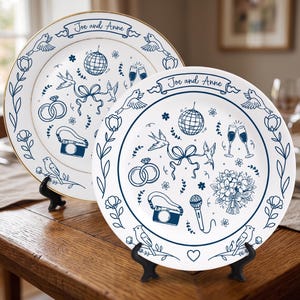May include: Two white ceramic plates with gold rims featuring blue line art illustrations of birds, flowers, wedding rings, a disco ball, a camera, a microphone, and a bouquet. The text "Joe and Anne" is printed on a banner at the top.