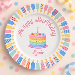 May include: A white ceramic plate featuring a colourful illustration of a multi-layered rainbow cake with a single lit candle in the centre. The text "Happy Birthday" and "Minnie" is printed in pink. The rim is decorated with various candles.