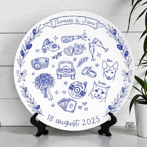 May include: A round white decorative plate featuring blue line art illustrations of a bouquet, a car, a record, a dog, a cat, and wedding symbols. The text reads Thomas & Jane at the top and 18 august 2025 at the bottom, framed by olive branches.