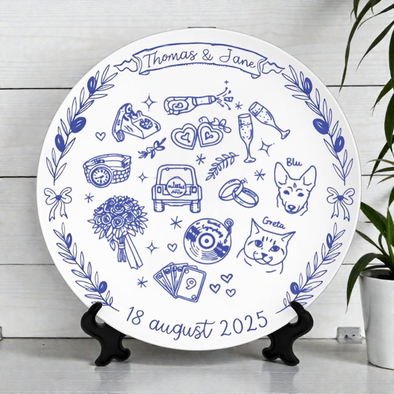 May include: A round white decorative plate featuring blue line art illustrations of a bouquet, a car, a record, a dog, a cat, and wedding symbols. The text reads Thomas & Jane at the top and 18 august 2025 at the bottom, framed by olive branches.