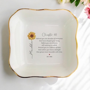 Può includere: Un piattino quadrato in ceramica bianca con un bordo smerlato dipinto in oro. Il centro presenta un'illustrazione di un girasole giallo, il nome Maddie e il testo: Chapter 60, Behind you are decades of memories, Before you are dreams, EST. 1966.