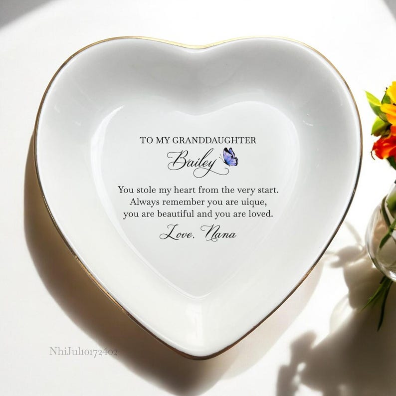 Puede incluir: Un plato blanco en forma de coraz&oacute;n con un borde dorado. El plato tiene un mensaje que dice: "A mi nieta Bailey Me robaste el coraz&oacute;n desde el principio. Recuerda siempre que eres &uacute;nica, eres hermosa y eres amada. Con amor. Abuela" con un gr&aacute;fico de mariposa azul.