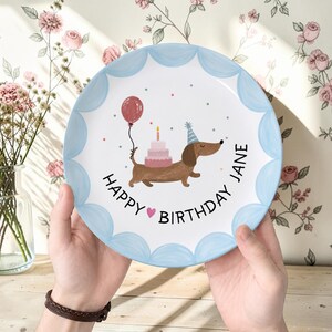 May include: A round ceramic plate with a scalloped light blue border featuring a brown dachshund wearing a party hat, a pink tiered birthday cake, a red balloon, and the text "HAPPY BIRTHDAY JANE" with a small pink heart in the center.