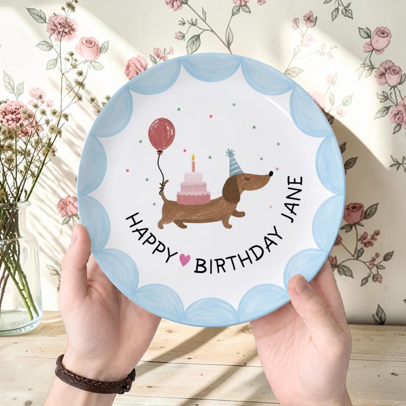 May include: A round ceramic plate with a scalloped light blue border featuring a brown dachshund wearing a party hat, a pink tiered birthday cake, a red balloon, and the text "HAPPY BIRTHDAY JANE" with a small pink heart in the center.