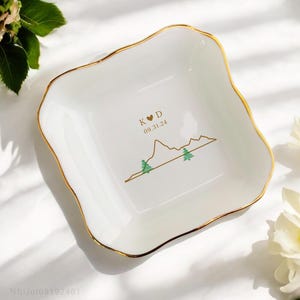 May include: A white ceramic trinket dish with a gold rim. The dish features a mountain range design with two pine trees and the text "K ♥ D 09.31.24".