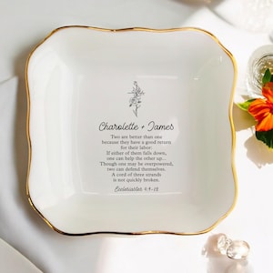 May include: A square white ceramic trinket dish with scalloped edges and a gold-painted rim. The centre features a black line drawing of a rose and cross, the names Charolette + James, and the text of Ecclesiastes 4:9-12 in black lettering.