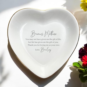 Op de afbeelding: Een hartvormige witte keramische sieradenschaal met een gouden rand. De schaal heeft de woorden "Bonus Mother" en een boodschap van waardering in zwart schrift. De boodschap is ondertekend met "Love, Bailey."