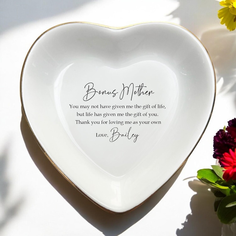 Op de afbeelding: Een hartvormige witte keramische sieradenschaal met een gouden rand. De schaal heeft de woorden "Bonus Mother" en een boodschap van waardering in zwart schrift. De boodschap is ondertekend met "Love, Bailey."