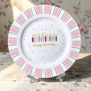 Puede incluir: Un plato de cerámica blanca con un borde de bloques rectangulares rosas alternados y líneas verdes finas. El centro muestra una fila de velas de cumpleaños coloridas con el texto happy birthday escrito en cursiva dorada.