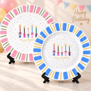 Pode incluir: Dois pratos redondos de cerâmica com centro branco, confetes coloridos, cinco velas de aniversário ilustradas e o texto Happy Birthday. O prato maior tem uma borda listrada em rosa e verde, enquanto o menor tem uma borda listrada em azul e amarelo.