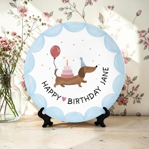 May include: A round ceramic plate with a scalloped light blue border featuring a brown dachshund wearing a blue party hat, carrying a pink tiered cake and a red balloon, with the text HAPPY BIRTHDAY JANE and a small pink heart printed.