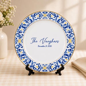 May include: A white ceramic plate with a gold rim and a blue and yellow floral scroll pattern along the border. The center features the blue text The Vaughars and the date December 21, 2026. The plate is displayed on a black plastic stand.