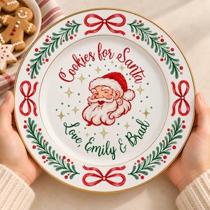 Puede incluir: Un plato de cerámica blanca con borde dorado que presenta una ilustración central de Santa Claus, el texto rojo "Cookies for Santa" y el texto verde "Love, Emily & Brad". El borde está decorado con lazos rojos y ramas de pino verdes.