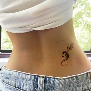 Pode incluir: Uma tatuagem de drag&atilde;o preto na parte inferior das costas de uma pessoa. O drag&atilde;o est&aacute; enroscado com as asas abertas. H&aacute; um pequeno ponto vermelho abaixo do drag&atilde;o.