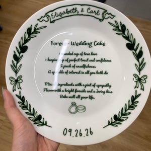 May include: A white ceramic plate featuring a dark green border of olive branches and bows. The center text reads Forever Wedding Cake with a recipe for love, the names Elizabeth & Carl, and the date 09.26.26, accompanied by two ring icons.