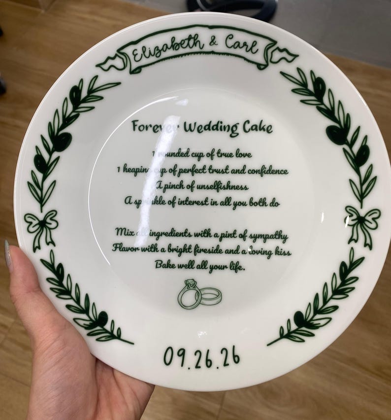 May include: A white ceramic plate featuring a dark green border of olive branches and bows. The center text reads Forever Wedding Cake with a recipe for love, the names Elizabeth & Carl, and the date 09.26.26, accompanied by two ring icons.