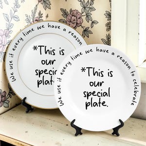 Puede incluir: Dos platos de cerámica blanca con bordes dorados, cada uno con texto escrito a mano en negro. El texto dice: *This is our special plate. We use it every time we have a reason to celebrate. Los platos se exhiben sobre soportes negros.