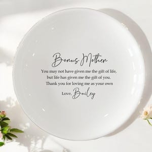 Op de afbeelding: Een witte keramische plaat met de woorden "Bonus Mother" in sierlijke letters. Daaronder staat een ontroerende boodschap en de naam "Bailey". De plaat is rond en heeft een gladde, glanzende afwerking.