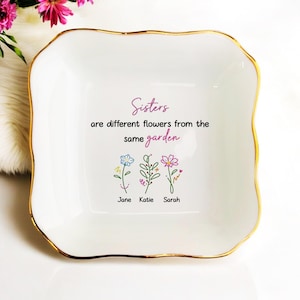 May include: A white ceramic trinket dish with a gold rim. The dish features the text "Sisters are different flowers from the same garden" and three illustrated flowers with the names Jane, Katie, and Sarah.