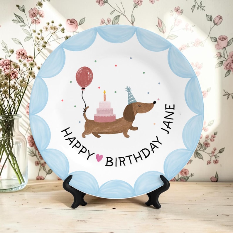May include: A white ceramic plate with a scalloped light blue border. The center features a brown dachshund wearing a blue party hat, carrying a pink tiered cake and a red balloon. The text reads Happy Birthday Jane with a small pink heart.