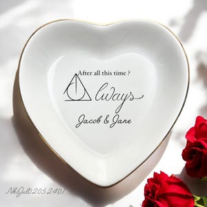 May include: White heart-shaped ceramic trinket dish with a gold rim. The dish features a black and gold Deathly Hallows symbol, the text "After all this time? Always", and the names "Jacob & Jane".