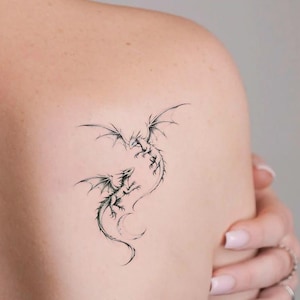 Könnte beinhalten: Zwei detaillierte schwarze Drachentattoos sind auf der Schulter einer Person abgebildet. Die Drachen blicken einander an, mit ausgebreiteten Flügeln und komplizierten Schuppen. Das Tattoo-Design ist im Fine-Line-Art-Stil.