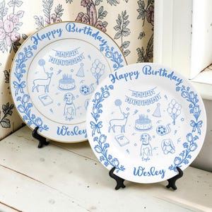 May include: Two white ceramic plates with blue line art illustrations of a deer, a dog, a birthday cake, balloons, and a banner reading Happy Birthday. The plates feature the names Wesley and Layla in blue script text along the bottom edge.