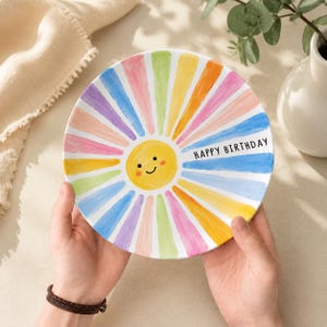 May include: A round ceramic plate featuring a central yellow sun with a smiling face, surrounded by radiating colourful painted stripes in shades of pink, orange, yellow, green, and blue. The text HAPPY BIRTHDAY is printed in black on the right side.