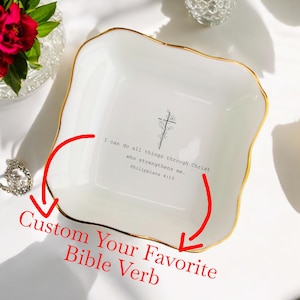 Op de afbeelding: Een wit keramiek sieradenschoteltje met een gouden rand. Het schoteltje heeft een kruisontwerp en de tekst "Ik kan alles door Christus, die mij kracht geeft. Filippenzen 4:13". De tekst "Maak je favoriete Bijbelvers" staat onder het schoteltje.