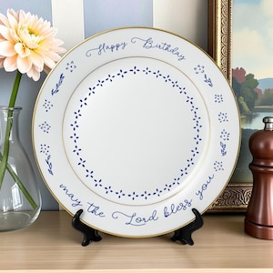 Pode incluir: Um prato de cerâmica branca com borda dourada, apresentando detalhes florais azuis e um padrão circular de pontos no centro. O texto "Happy Birthday" está impresso no topo e "may the Lord bless you" na parte inferior em azul.