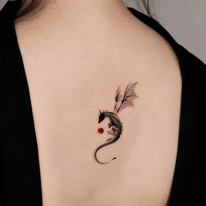 Pode incluir: Uma tatuagem de drag&atilde;o preto e cinza nas costas de uma pessoa. O drag&atilde;o est&aacute; enroscado com as asas abertas. H&aacute; um pequeno c&iacute;rculo vermelho atr&aacute;s do drag&atilde;o.