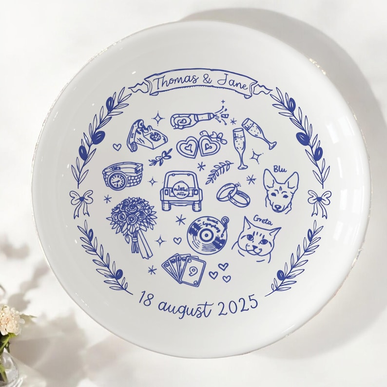May include: A white ceramic plate featuring blue line art illustrations of a wedding theme, including a car, rings, a bouquet, a record, a dog named Blu, and a cat named Greta. The text reads Thomas & Jane and 18 august 2025.