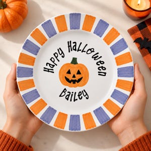 Może przedstawiać: Biały talerz ceramiczny z ręcznie malowaną pomarańczową dynią pośrodku z czarną twarzą jack-o'-lantern. Napis "Happy Halloween" oraz "Bailey" jest wydrukowany na czarno. Brzeg talerza ma naprzemienne pomarańczowe i fioletowe prostokątne paski.