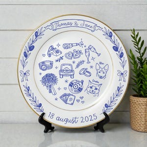 May include: A white ceramic plate with a gold rim featuring blue line art illustrations including a car, a bouquet, a vinyl record, playing cards, and pets named Blu and Greta. The text reads Thomas & Jane and 18 august 2025.