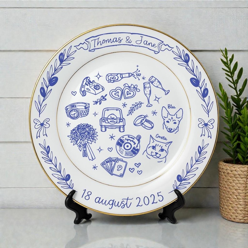 May include: A white ceramic plate with a gold rim featuring blue line art illustrations including a car, a bouquet, a vinyl record, playing cards, and pets named Blu and Greta. The text reads Thomas & Jane and 18 august 2025.
