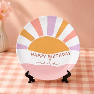 Könnte beinhalten: Ein runder Keramikteller mit einer handgemalten gelben Sonne und strahlenförmigen Streifen in Rosa, Lila und Gelb. Der Text "HAPPY BIRTHDAY" ist in Braun gedruckt, gefolgt von dem Namen "mila" in Schreibschrift über einer rosa Wellenlinie.