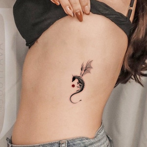 Pode incluir: Uma tatuagem de drag&atilde;o preto no lado de uma pessoa com um pequeno ponto vermelho abaixo.