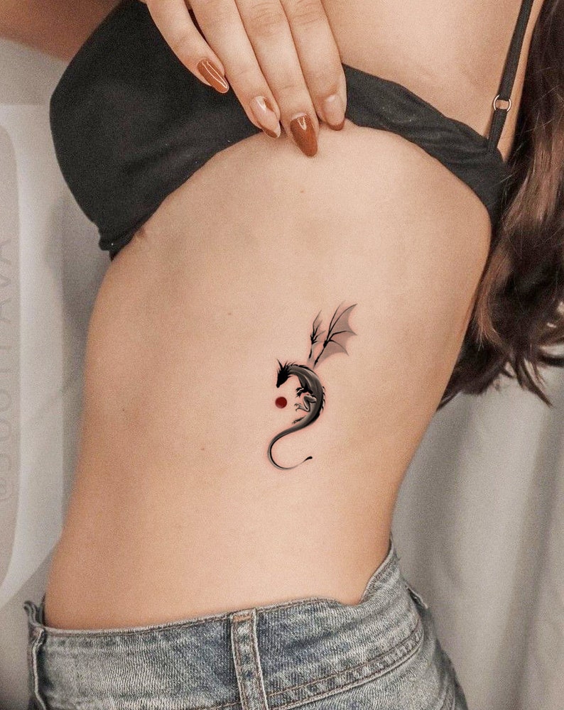 Pode incluir: Uma tatuagem de drag&atilde;o preto no lado de uma pessoa com um pequeno ponto vermelho abaixo.
