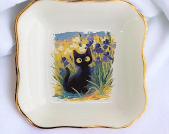 Pode incluir: Um prato de cerâmica branca com borda dourada apresenta um gato preto com olhos amarelos sentado em um campo de flores amarelas e roxas.