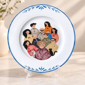 Puede incluir: Un plato de cerámica blanca con un borde floral pintado en azul que presenta una ilustración central de un grupo de personas con jerséis con las letras H, M, A, S, J y E, un gato negro pequeño y una persona con una camisa con las letras TC.