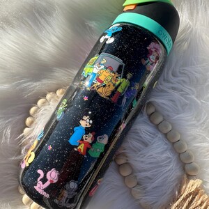 May include: Black water bottle with a variety of cartoon character decals, including Scooby-Doo, The Smurfs, and Alvin and the Chipmunks. The bottle has a black lid with a green band and a yellow top. The bottle is covered in glitter.
