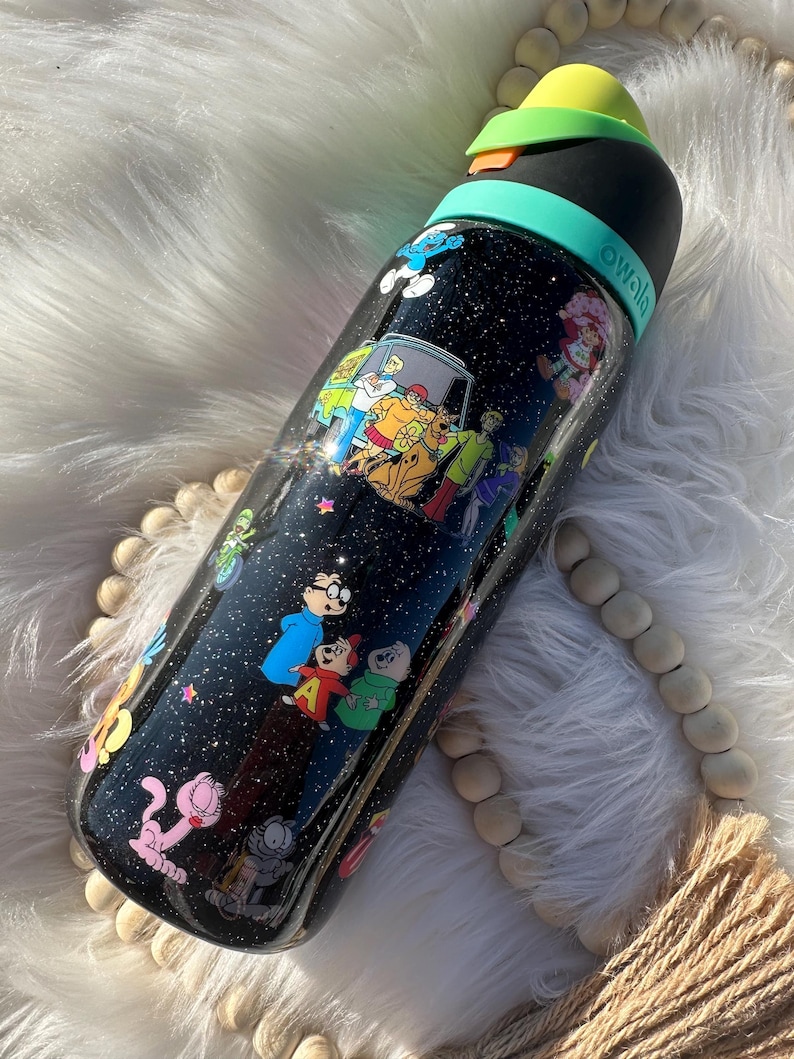 May include: Black water bottle with a variety of cartoon character decals, including Scooby-Doo, The Smurfs, and Alvin and the Chipmunks. The bottle has a black lid with a green band and a yellow top. The bottle is covered in glitter.