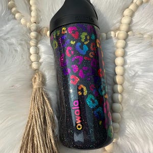 Op de afbeelding: Een zwarte Owala waterfles met een zwarte deksel. De fles is bedekt met kleurrijke luipaardvlekken en glitter. Het Owala-logo is roze. De fles staat tegen een witte, zachte achtergrond.