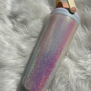 May include: A pink and white iridescent water bottle with a light blue lid and a yellow handle. The bottle has a pink logo that says "Owolo" on the side.