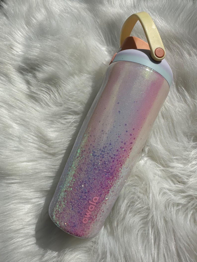 May include: A pink and white iridescent water bottle with a light blue lid and a yellow handle. The bottle has a pink logo that says "Owolo" on the side.