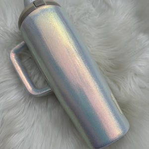 Puede incluir: Botella de agua iridiscente con asa. La botella tiene un acabado nacarado con tapa y asa de color gris claro. El botón pulsador es visible en la tapa. La botella está sobre una superficie blanca y esponjosa.