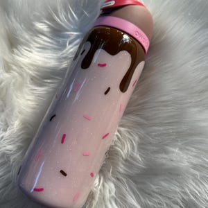 Puede incluir: Una botella de agua rosa brillante con una parte superior y goteo de color chocolate, que se asemeja a un helado. La botella tiene chispas rosas y marrones y una banda rosa alrededor de la parte superior. La tapa es blanca y tiene una banda roja.