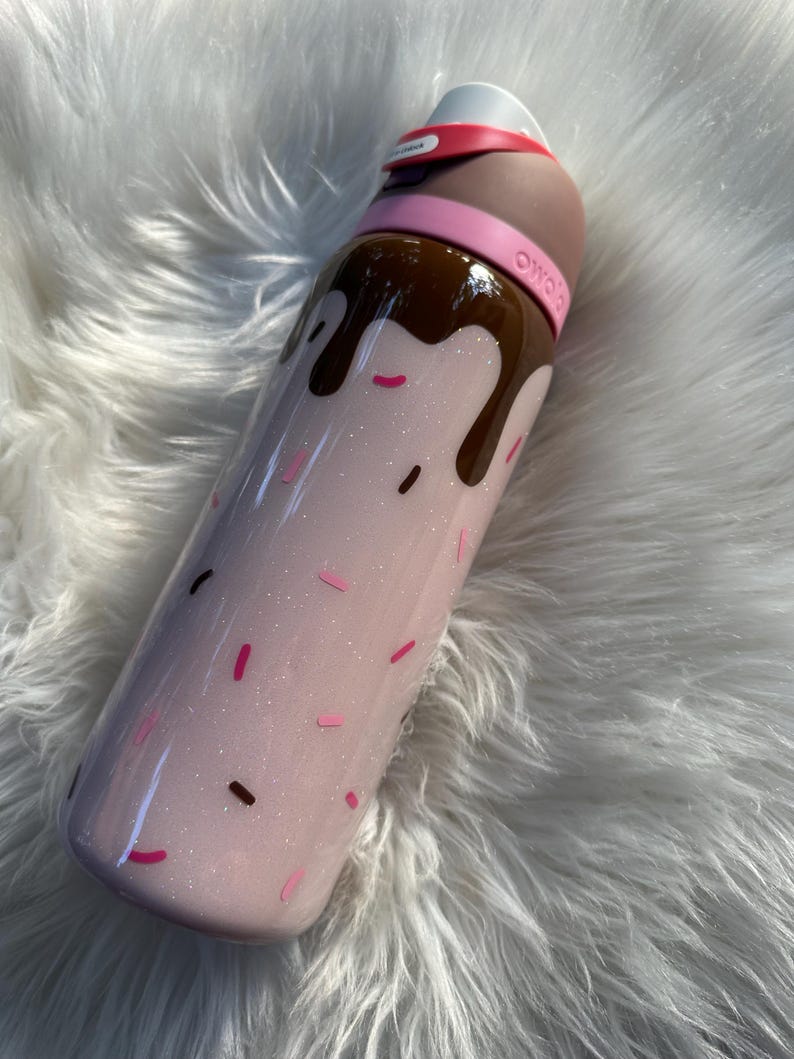 Puede incluir: Una botella de agua rosa brillante con una parte superior y goteo de color chocolate, que se asemeja a un helado. La botella tiene chispas rosas y marrones y una banda rosa alrededor de la parte superior. La tapa es blanca y tiene una banda roja.