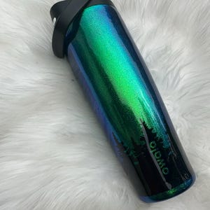 Peut inclure: Une gourde haute et irisée avec une poignée et un couvercle noirs. La bouteille passe du vert au bleu avec un motif de silhouette de forêt et le mot "OWOLO" imprimé sur le côté. La bouteille est sur une surface blanche et duveteuse.