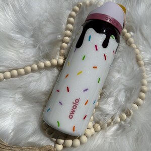 Puede incluir: Botella de agua reutilizable blanca con tapa rosa y un dise&ntilde;o marr&oacute;n parecido al chocolate con chispas de colores. La botella tiene la palabra "owala." impresa en ella.